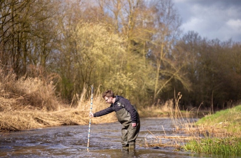 een collega staat in een waadpak in een beek en meet de waterkwaliteit